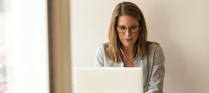 Two women standing and one woman sitting looking at a computer screen with bookshelves and an American flag in the background.